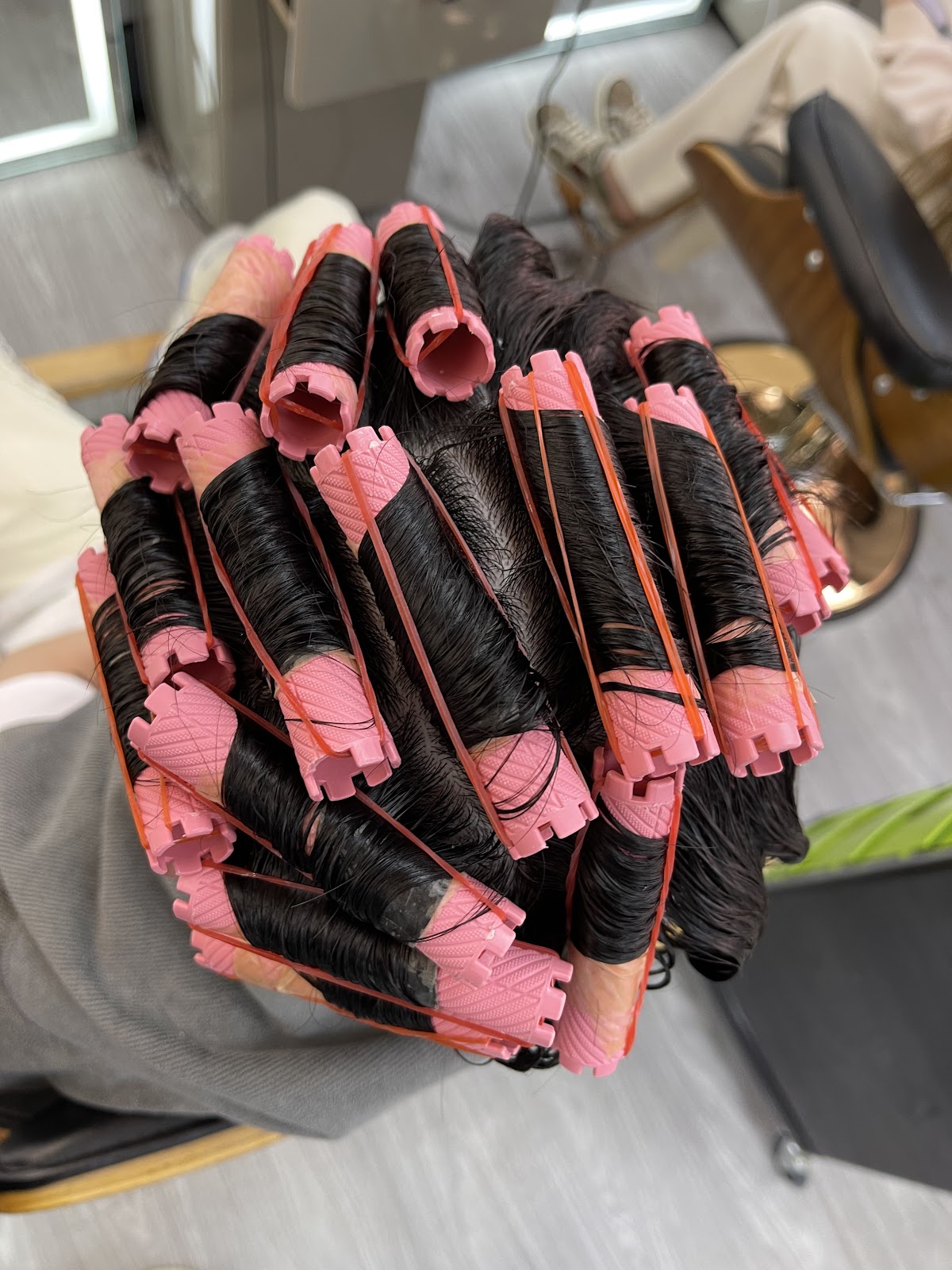 A head of dark hair wound onto pink perm rods held with orange elastics, seen from above.