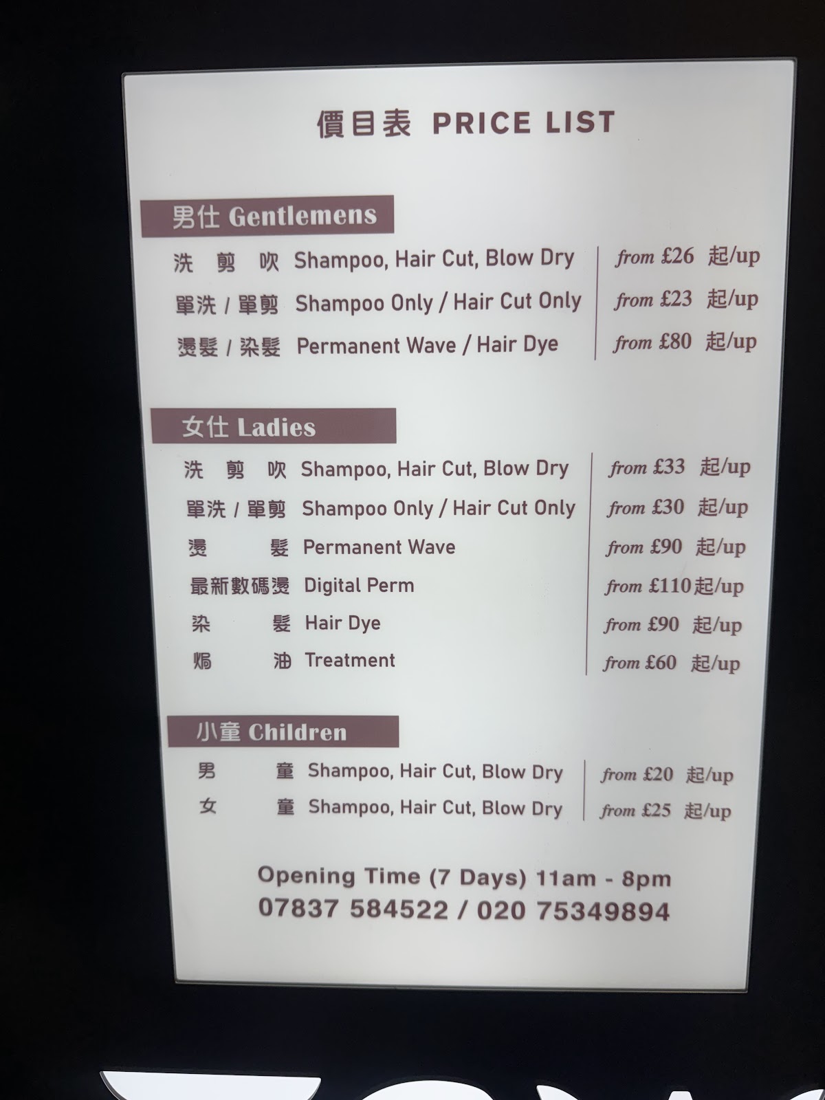 The salon's illuminated price board: a white panel headed 價目表 PRICE LIST, with Gentlemens, Ladies and Children sections in Chinese and English, and the opening time and two phone numbers along the bottom.