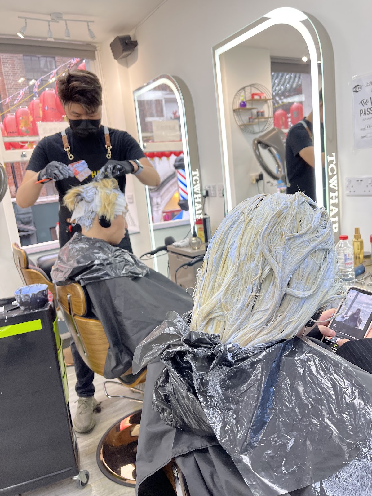 A stylist in a black apron applying bleach to a seated client's hair, in front of an arched, light-rimmed mirror carrying a TCW HAIR decal; red lanterns and bunting hang in the window behind.