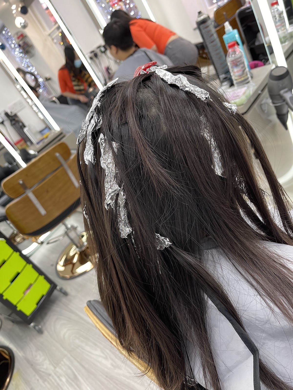 Foils being placed at the roots of long dark hair, with other clients in chairs at the lit mirrors behind.