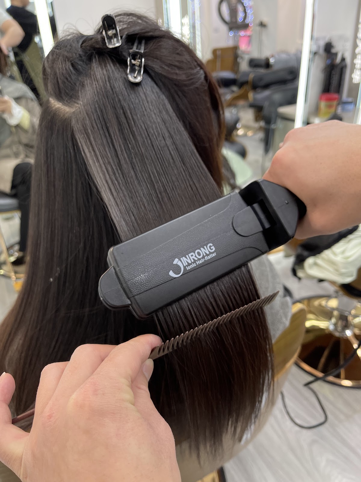 A stylist running a black ionic flat-iron and a comb through a long, glossy dark-brown length of hair.